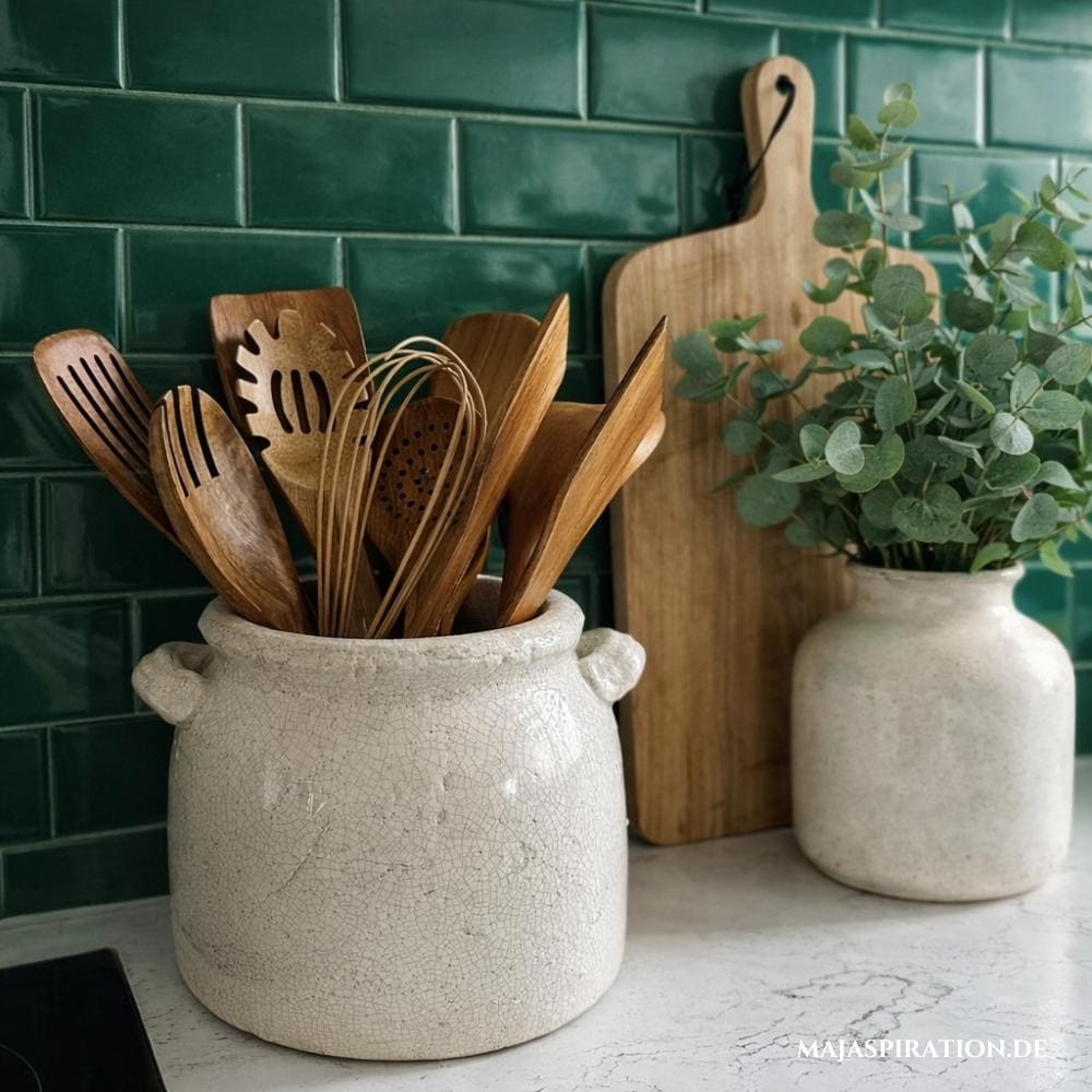 Küchenutensilien aus Holz in Keramiktopf vor grünen Küchenfliesen