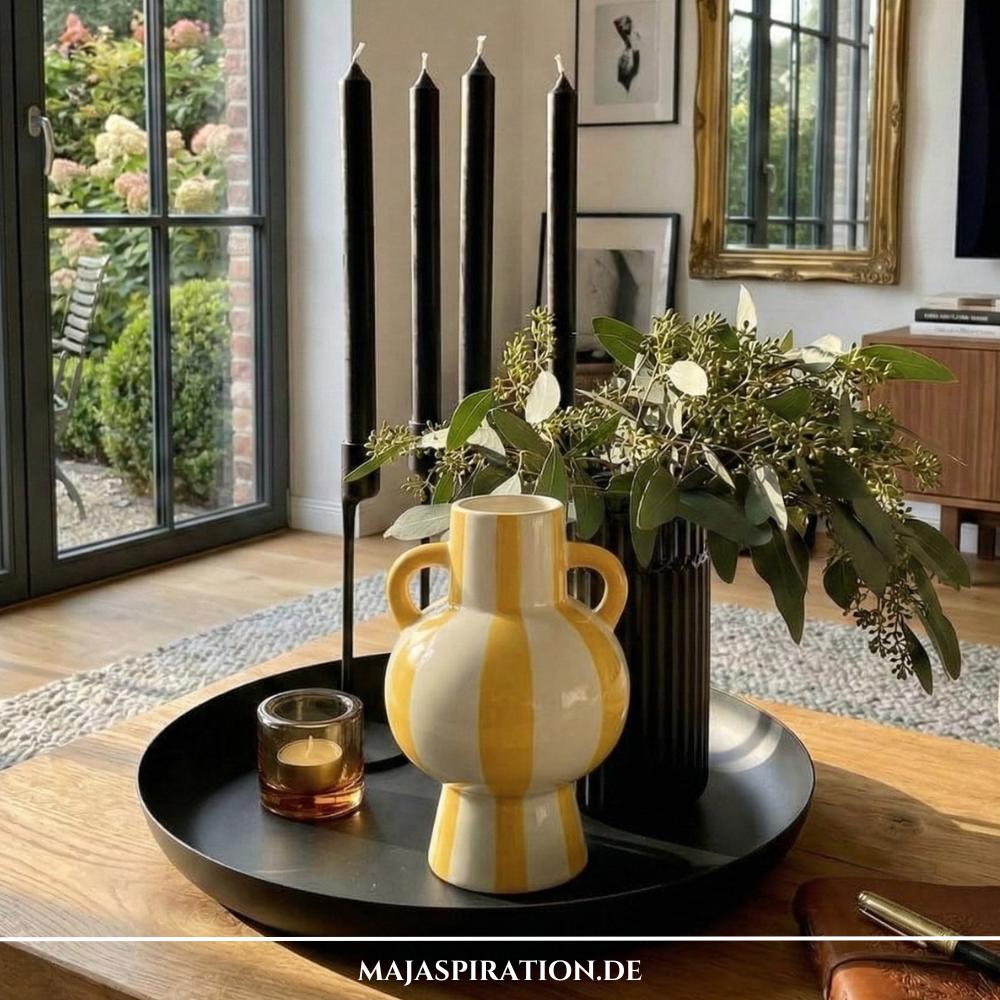 Rundes schwarzes Metalltablett mit schwarzen Stabkerzen, gelber Vase und Eukalyptus als moderne Tablett Deko auf einem hölzernen Couchtisch.