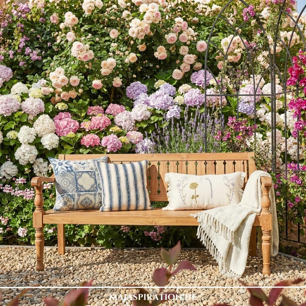 Romantische Holzbank im Landhaus-Stil vor einer üppigen Wand aus Hortensien und Rosen, dekoriert mit blau-weißen Kissen und einer Wolldecke – ein Beispiel für die naturnahen Gartentrends 2026.