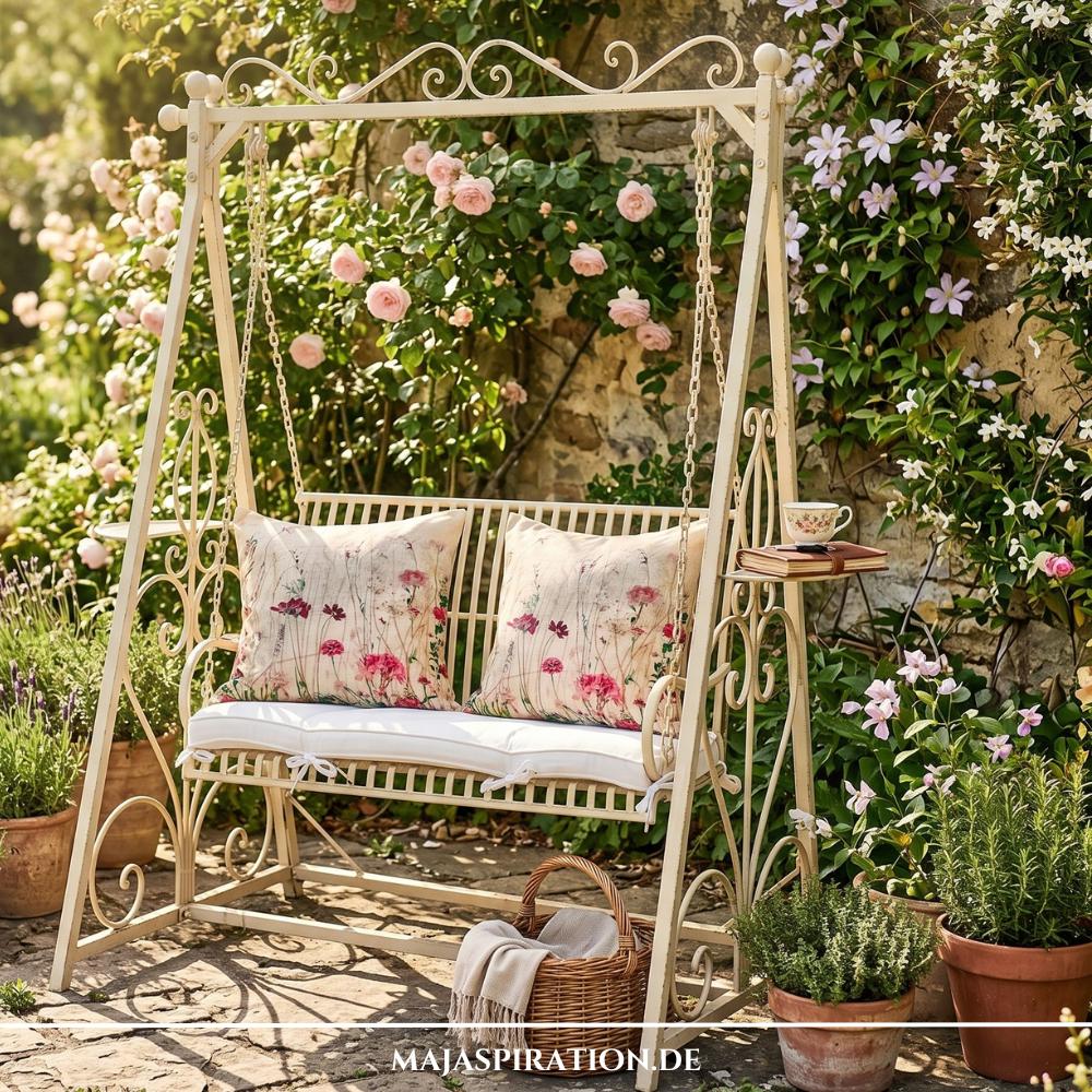 Romantische Hollywoodschaukel aus cremefarbenem Metall im Landhausstil vor einer Rosenwand, repräsentativ für die Gartentrends 2026 im Bereich Vintage-Gartenmöbel und Cottage-Design.