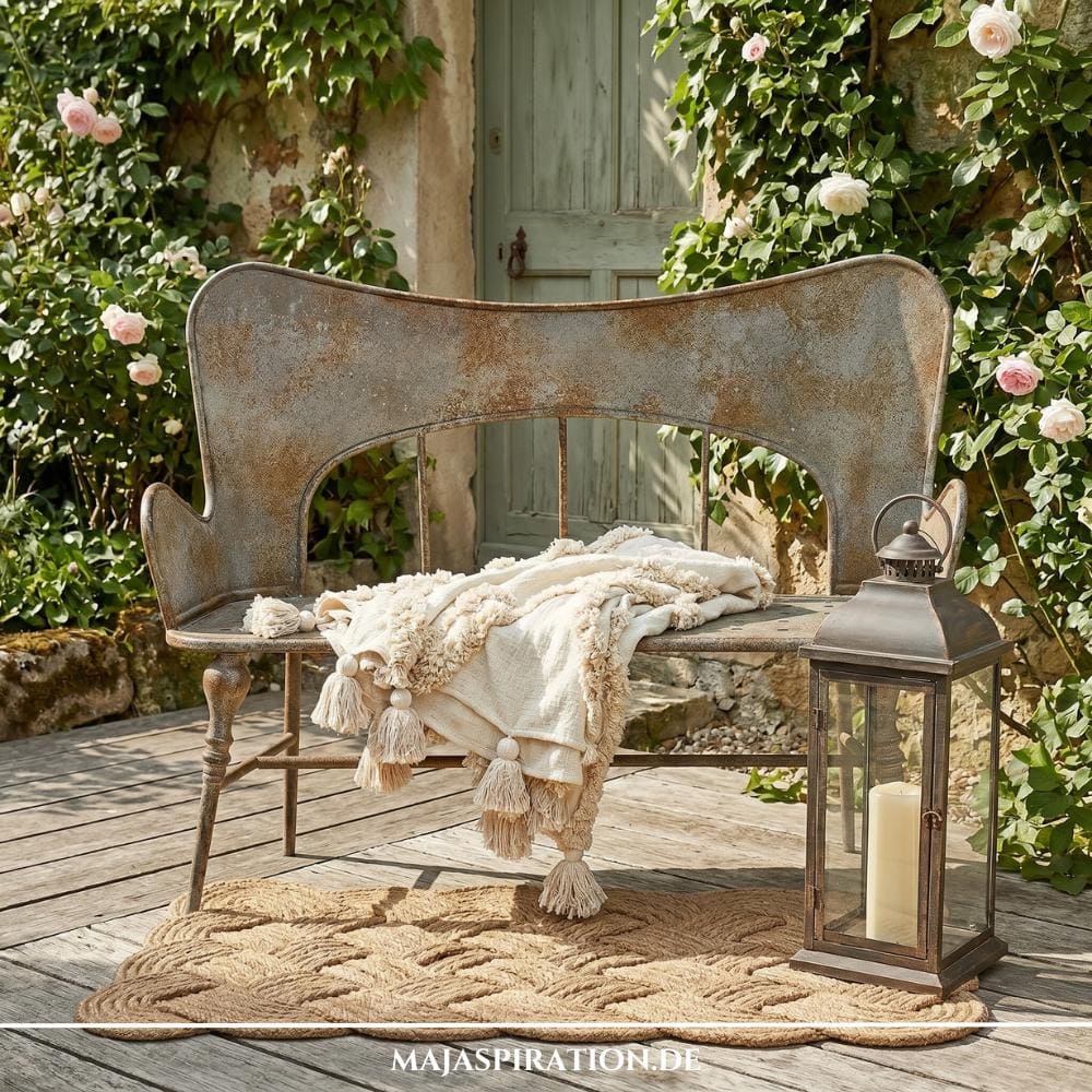 Romantische Gartenbank aus Metall mit Patina im Shabby-Look, dekoriert mit Boho-Decke und Laterne auf Holzterrasse – ein Beispiel für die naturnahen Gartentrends 2026 im Landhausstil.