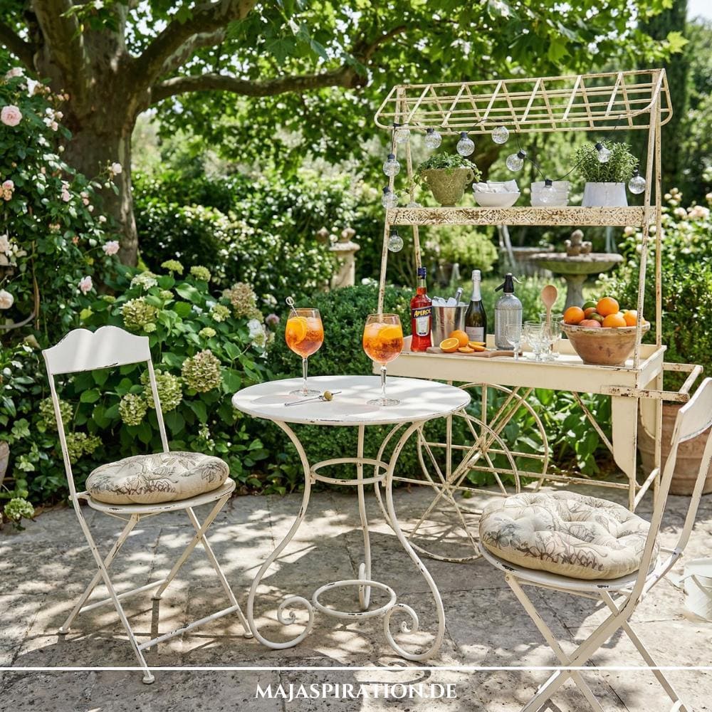 Weißes Metall-Bistro-Set und ein dekorativer Garten-Servierwagen mit Getränken und Lichterkette. Romantische Gestaltungsidee für die Gartentrends 2026 von MAJASPIRATION.DE.