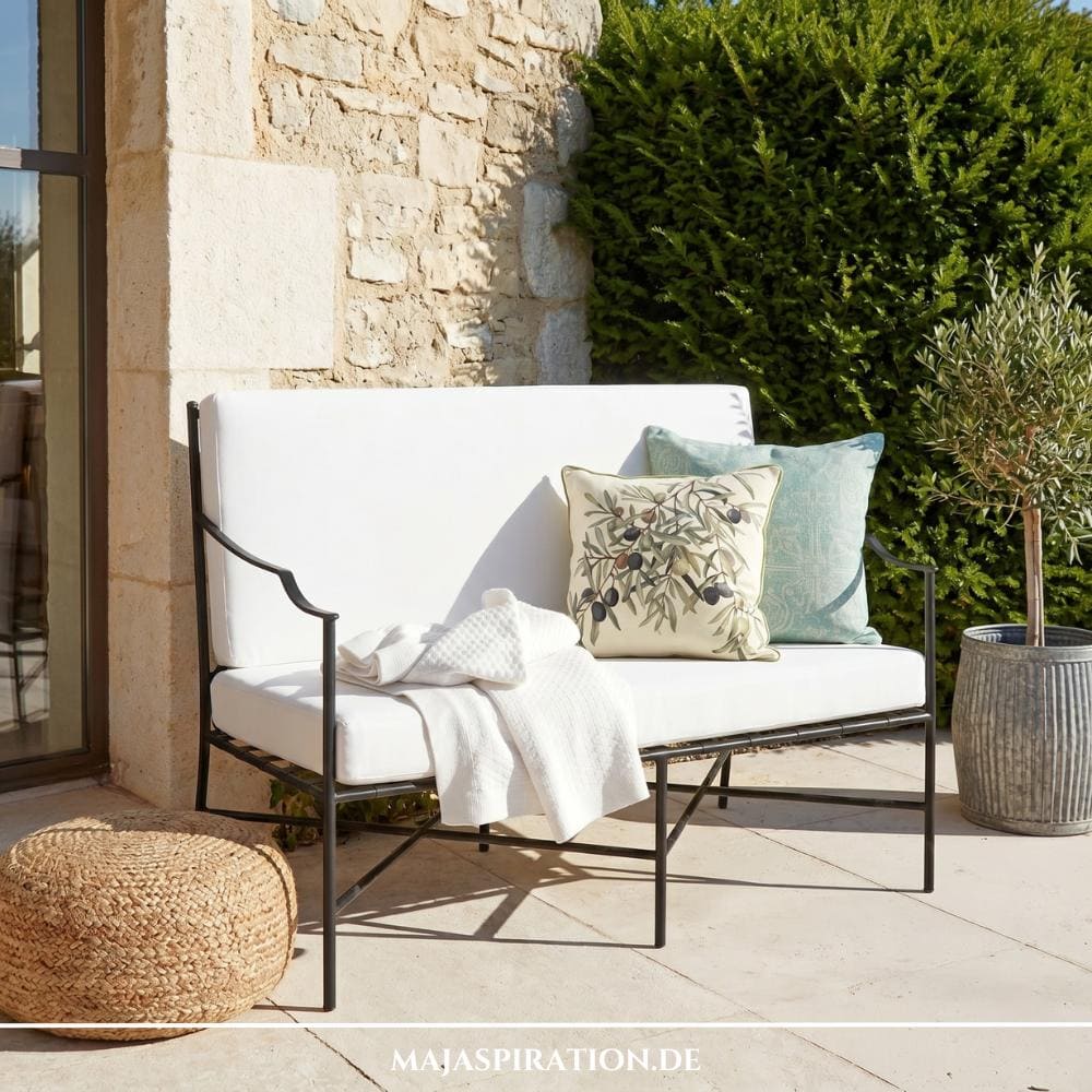 Metall-Sitzbank im romantischen Landhausstil mit weißen Polstern und Dekokissen vor einer Natursteinwand, ein Beispiel für die naturnahen Gartentrends 2026.