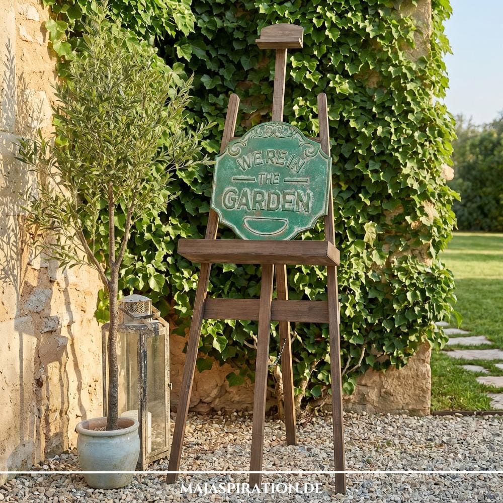 Kreative Cottage-Gartengestaltung im Sinne der Gartentrends 2026, mit einer rustikalen Holz-Staffelei, einem Vintage-Metallschild und einem Olivenbaum vor einer Efeumauer.