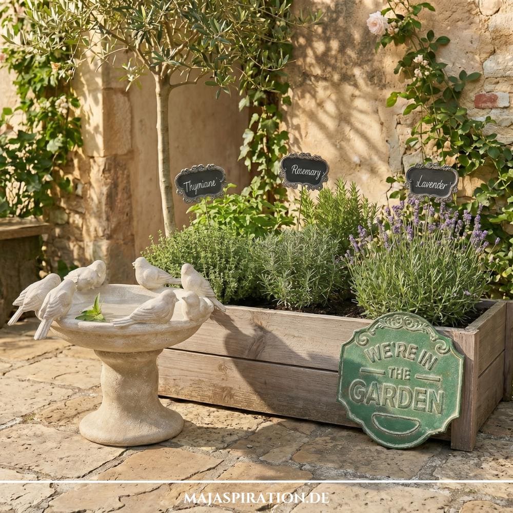 Romantische Gartentrends 2026: Eine klassische Stein-Vogeltränke neben einem rustikalen Holz-Kräuterbeet mit Lavendel, Rosmarin und Thymian vor einer mediterranen Natursteinwand im Cottage-Stil.
