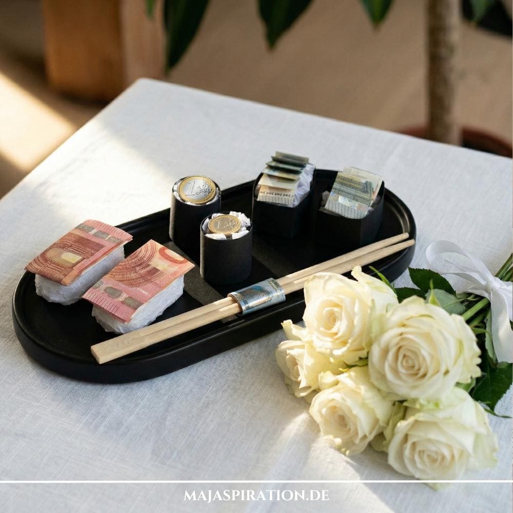 Geldgeschenk Reisekasse: Kreativ gefaltete Euro-Scheine und Münzen als Sushi-Set auf einem schwarzen Tablett mit Essstäbchen und weißen Rosen als Hochzeitsgeschenk.