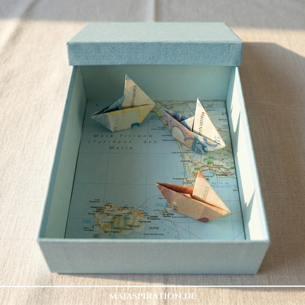 DIY Reise-Geldgeschenk: Drei aus Geldscheinen gefaltete Origami-Schiffe in einer hellblauen Box mit einer italienischen Landkarte (Ischia & Pozzuoli) als Hintergrund – kreative Idee für die Urlaubskasse und Flitterwochen.