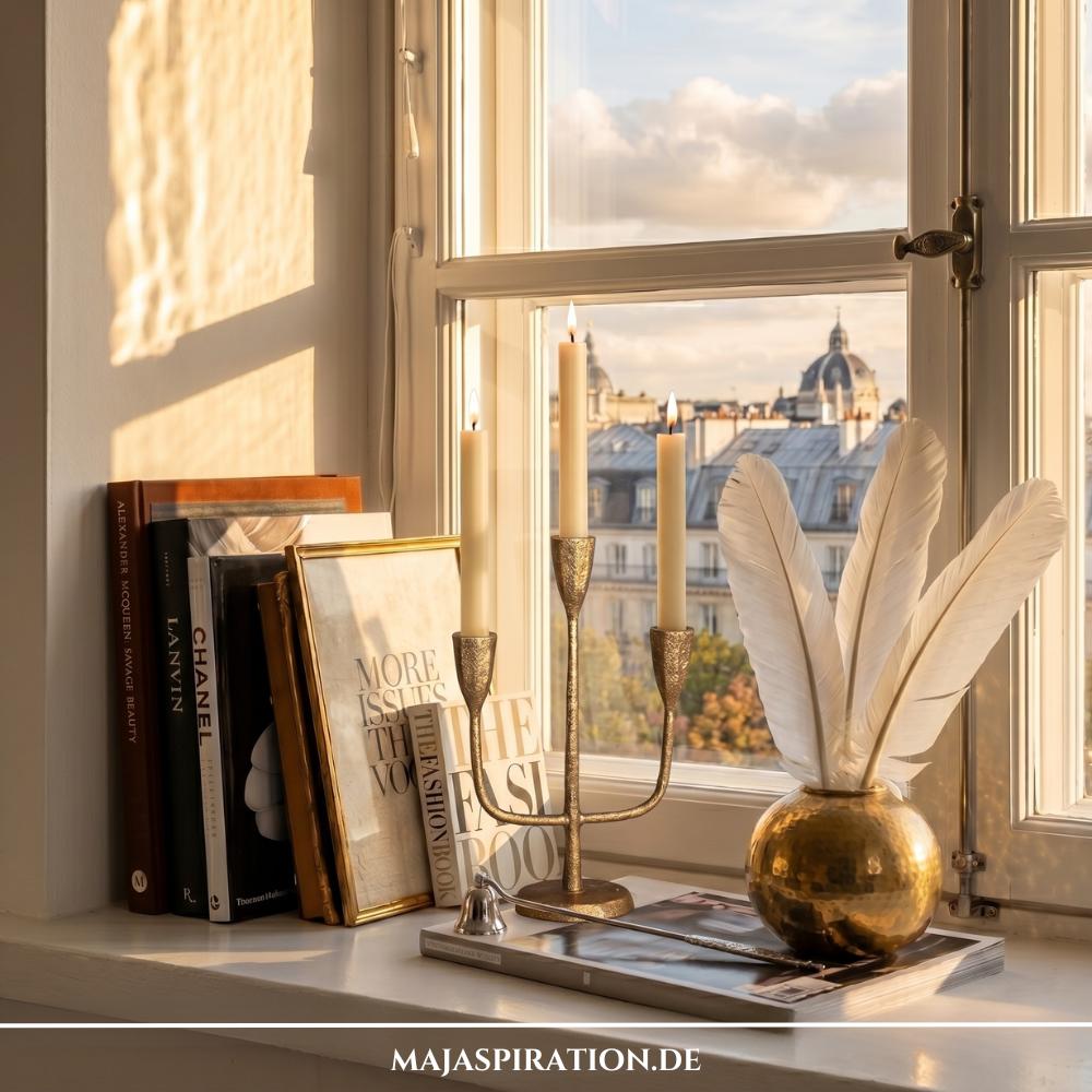 Fensterbank Deko Idee im Pariser Vintage-Chic bei warmem Abendlicht vor einem Fenster mit Blick auf D&auml;cher mit Kuppelgeb&auml;uden. Auf der wei&szlig;en Fensterbank lehnen links gro&szlig;e Mode-Bildb&auml;nde und gerahmte Typografie-Poster in Goldrahmen an der Wand. In der Mitte steht ein mehrarmiger, antiker Kerzenleuchter aus Messing mit gelb-braunen Kerzen auf einem flachen Buch, w&auml;hrend rechts eine runde, geh&auml;mmerte Goldvase mit Eukalyptuszweigen und einer wei&szlig;en Feder das elegante Bild abrundet.