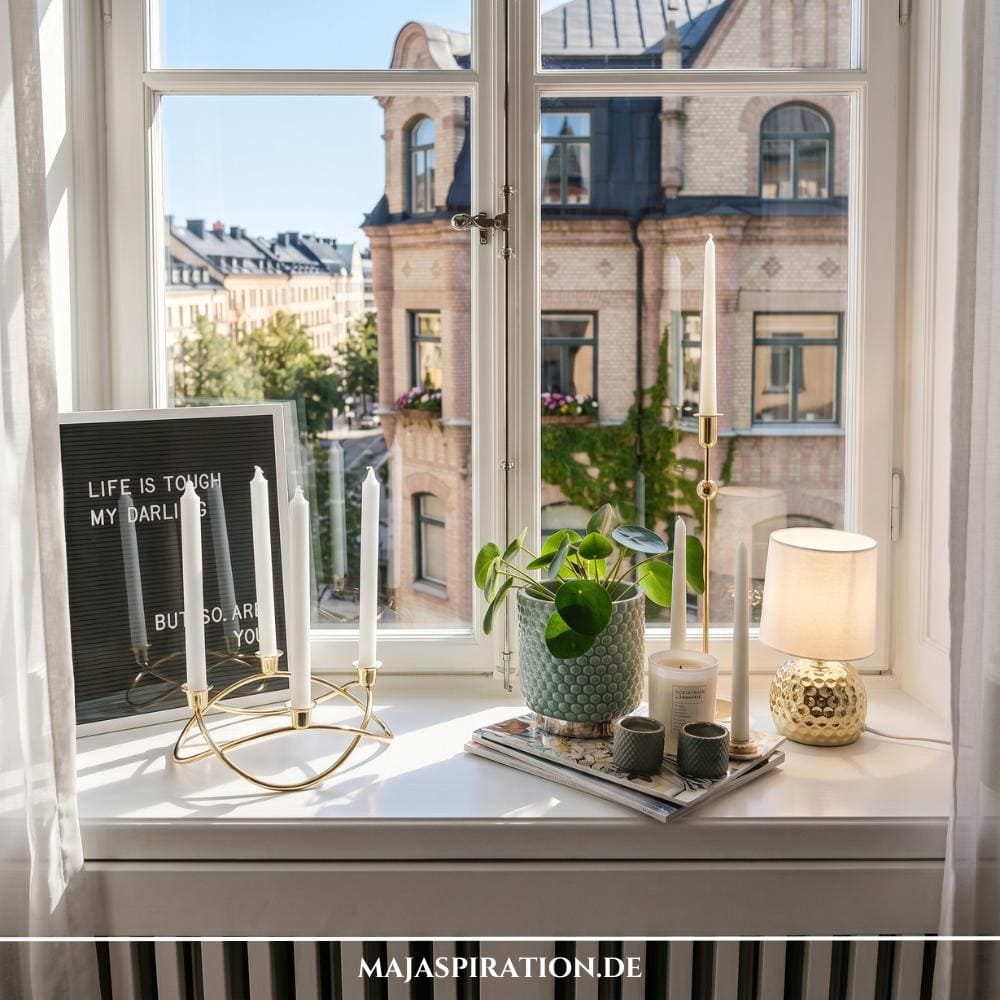 Sonnendurchflutete, wei&szlig;e Fensterbank im skandinavischen Design vor einem hellen Sprossenfenster mit Blick auf historische Backsteingeb&auml;ude. Auf der linken Seite lehnt ein schwarzes Letterboard mit wei&szlig;er Schrift an der Wand, direkt hinter einem bogenf&ouml;rmig geschwungenen, goldenen Kerzenst&auml;nder mit vier wei&szlig;en Stabkerzen. Mittig steht eine gr&uuml;ne Pilea-Pflanze in einem gemusterten Tontopf auf Magazinen, umgeben von kleinen Windlichtern und einer filigranen, hohen Kerze. Eine kleine Tischleuchte mit gl&auml;nzendem, goldenem Relief-Fu&szlig; und hellem Stoffschirm schlie&szlig;t das Arrangement rechts ab.