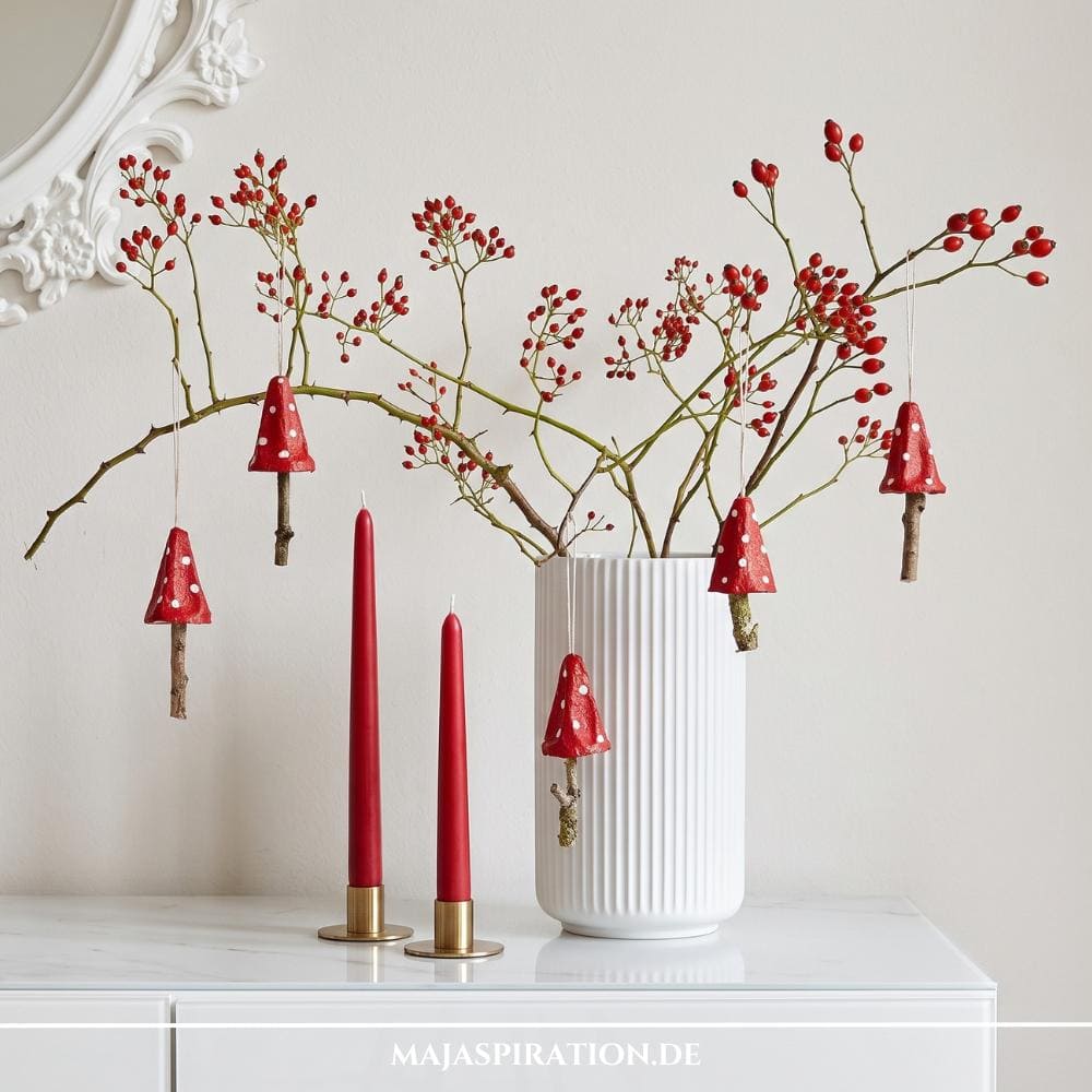 Vier rote, handgemachte DIY-Fliegenpilz-Anhänger mit weißen Punkten und Holzstielen, hängend an Hagebuttenzweigen in einer weißen Rillenvase neben goldenen Kerzenständern mit roten Stabkerzen als elegante Herbstdeko.