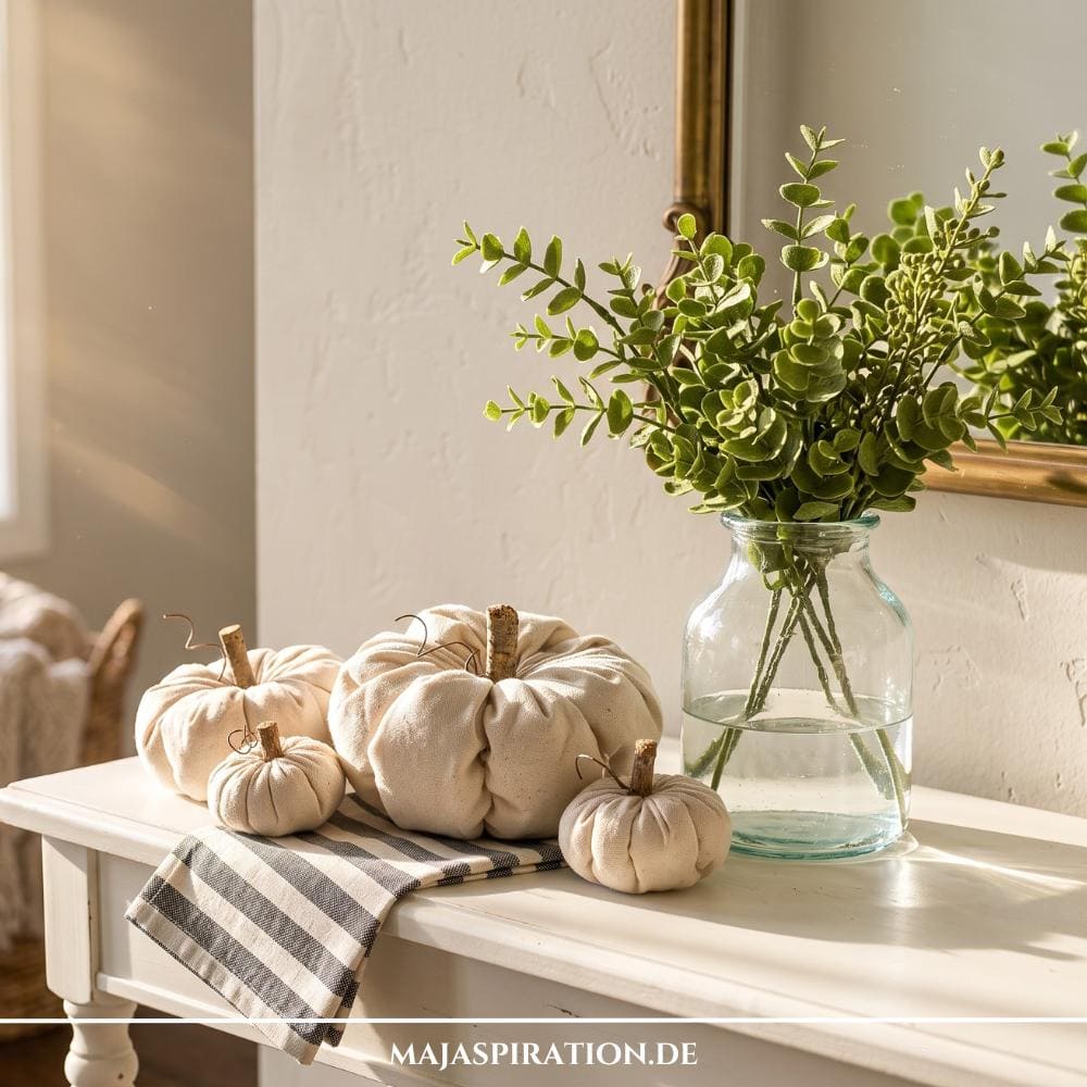 Vier cremefarbene DIY-Stoffkürbisse aus Leinen mit kleinen Holzstielen arrangiert auf einer hellen Konsole neben einer Glasvase mit grünen Eukalyptuszweigen für eine minimalistische, nachhaltige Herbstdeko im Cozy-Home-Stil.