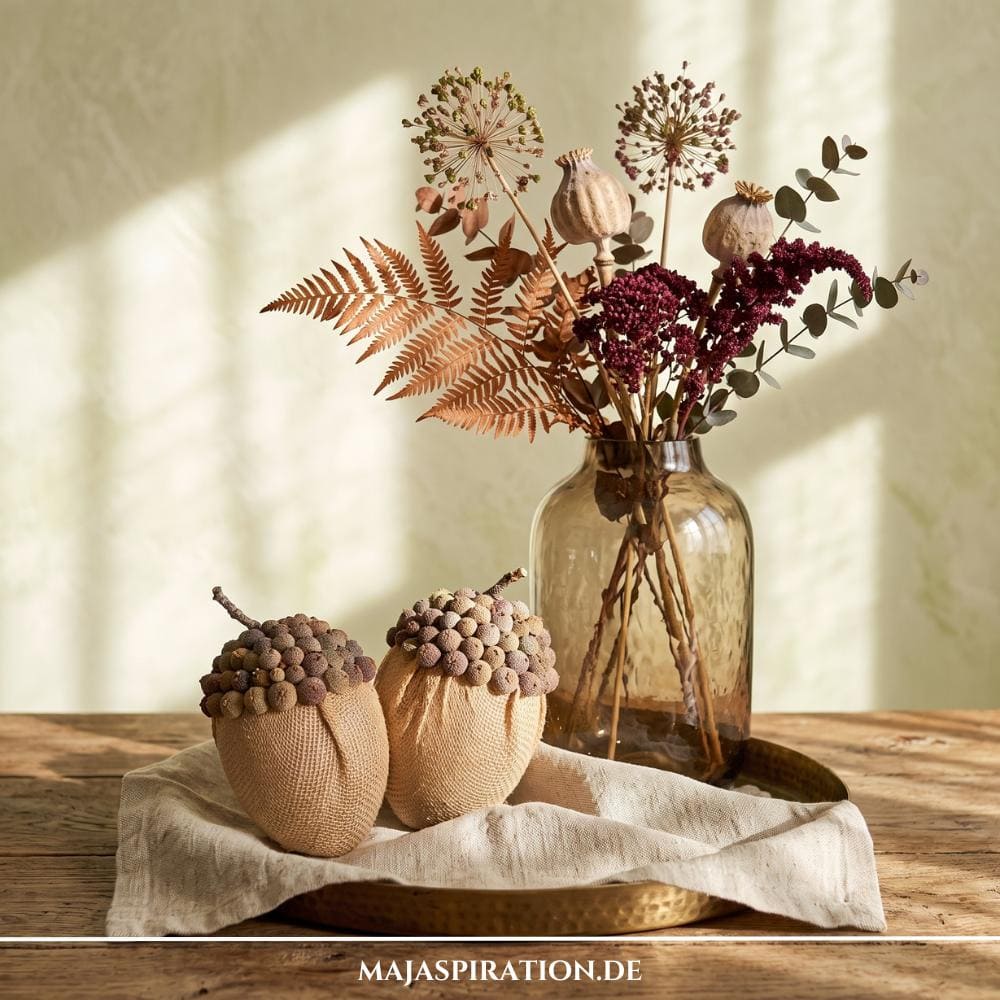Zwei große, handgemachte DIY-Deko-Eicheln aus beigem Jutestoff mit Kappen aus echten Erlenzapfen, arrangiert auf einem goldenen Tablett neben einer braunen Glasvase mit herbstlichen Trockenblumen auf einem Holztisch.