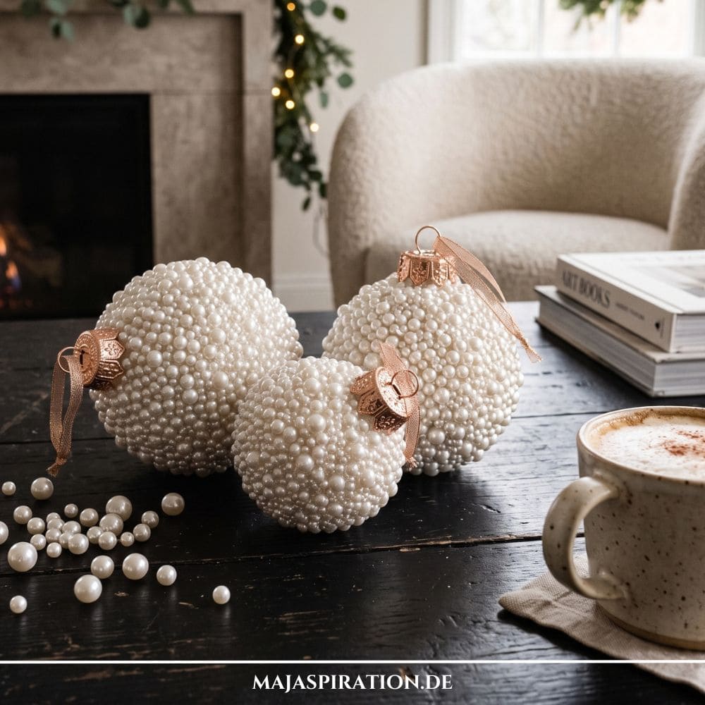 Nat&uuml;rliche DIY Weihnachtsdeko: Drei kunstvoll mit schimmernden cremewei&szlig;en Perlen verzierte Weihnachtskugeln auf einem dunklen Holztisch neben einer Tasse Kaffee.