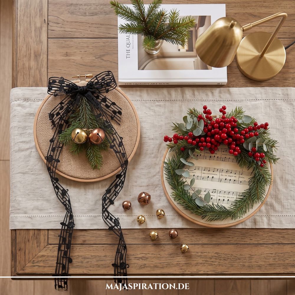 Rustikale Weihnachtsdekoration auf einem Holztisch mit Tischl&auml;ufer. Links liegt ein Stickrahmen mit Jute, einem Kiefernzweig, Glocken und einer Schleife aus Notenband; rechts ein Stickrahmen mit einem Kranz aus roten Beeren und Eukalyptus &uuml;ber Notenpapier.
