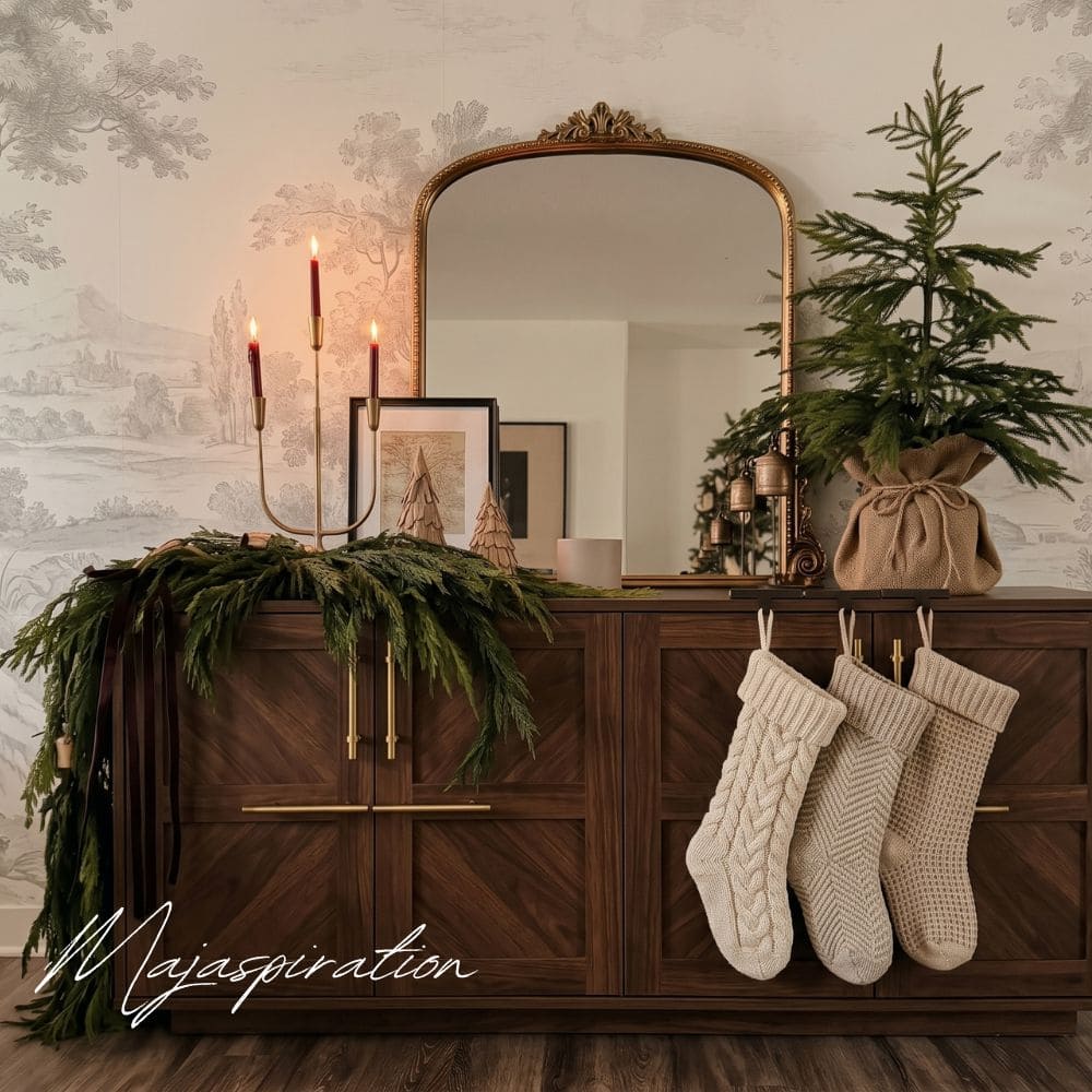 Weihnachtlich dekorierter dunkler Holz-Konsolentisch mit einem goldfarbenen Bogenwandspiegel, drei cremefarbenen Strickstr&uuml;mpfen, einem kleinen Nadelbaum im Jutesack und brennenden roten Kerzen auf einem Messingkerzenst&auml;nder vor einer Toile-de-Jouy-Tapete.