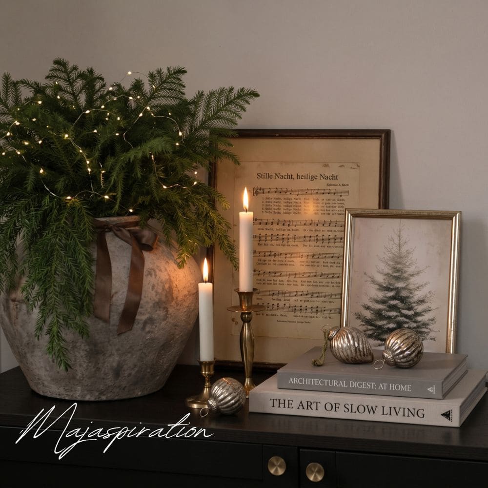 Weihnachtsdeko auf einem Konsolentisch mit beleuchtetem Tannengr&uuml;n in einer Steinvase, zwei Messingkerzenst&auml;ndern, Vintage-Weihnachtskugeln auf Coffee Table Books und eingerahmten Noten von &bdquo;Stille Nacht, heilige Nacht&ldquo;.