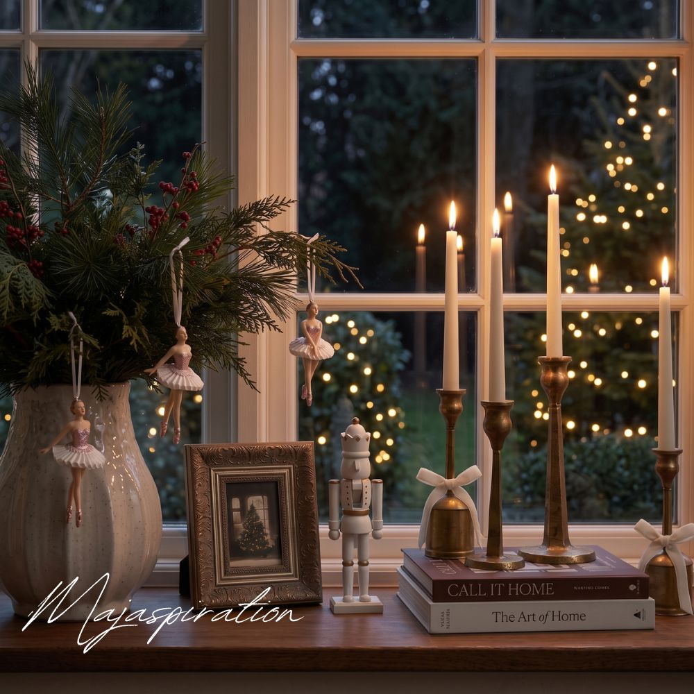 Festlich dekorierte Holzfensterbank zur Weihnachtszeit mit warm brennenden Stabkerzen in Messinghaltern auf Bildb&auml;nden, einer Vase mit Kiefernzweigen und Ballerina-Anh&auml;ngern sowie einem wei&szlig;en Nussknacker vor dem erleuchteten Abendfenster.